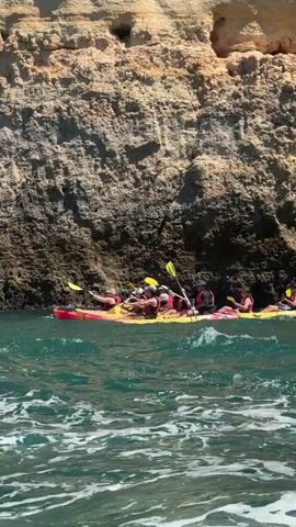 The video shows a group of people kayaking in the ocean near a rocky cliff face. The sun is shining brightly, indicating it's a clear day, likely daytime. The water is a vibrant turquoise and appears to be somewhat choppy with whitecaps. The cliffs are a light tan color with darker, textured rock formations and some vegetation growing at the top. Several kayaks, each with two people wearing life vests, are paddling close to the cliffs. The kayaks are primarily yellow and red. The people appear to be enjoying the activity. The footage pans to reveal more of the coastline and the kayaks navigating through the water. The overall impression is a scenic outdoor adventure in a coastal environment. No visible text is present in the video.