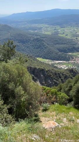 The video shows a panoramic view of a mountainous landscape. In the foreground, there's a rocky cliff partially covered with lush green vegetation. Below the cliff, a dense forest extends towards a valley. The valley contains a scattering of buildings, possibly a small town or village, nestled amidst fields and a winding road. Beyond the valley, the terrain transitions into flatter, cultivated land, dotted with agricultural fields. In the distance, more mountains rise, their peaks obscured by a hazy blue sky. The scene is bathed in sunlight, suggesting a clear, bright day.