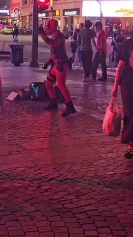 The video shows a person dressed in a Deadpool costume dancing on a red brick surface at night. The person is wearing a full Deadpool suit with red and black colors, along with dark boots. A small, lit-up speaker sits near the person, presumably playing music. There are pedestrians in the background, and a McDonald's restaurant and digital billboards are visible across the street. The video is filmed at night, with artificial lighting casting a reddish hue over the scene.