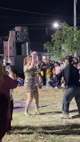 A woman in a sparkly gold dress stands center frame, holding a glass, and appears to be singing or performing for an audience. She is facing a man in a black shirt and jeans who is standing to her right. Behind them, a crowd of people are watching the performance. In the background, there are large speakers and a flag. The scene takes place outdoors at night.