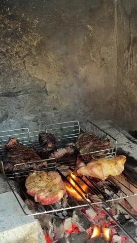 Several cuts of meat are arranged on a metal grill over burning coals inside what appears to be a stone fireplace. The meats vary in doneness, with some looking well-seared and others still pink.  Smoke rises from the coals.