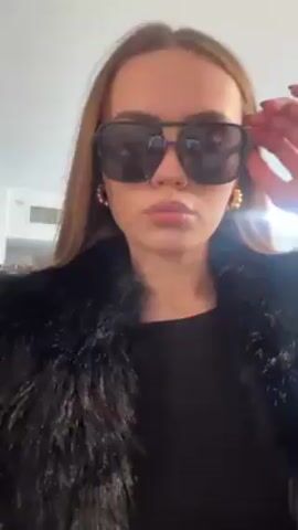 A woman with light brown hair and sunglasses is the focus of this video. She is wearing a black fur coat and a black top. She has hoop earrings. She adjusts her sunglasses. She puckers her lips. The background is blurry but appears to be an interior space.