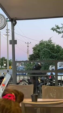 The camera is positioned inside an open-air café looking out onto a street at dusk. The sky is tinged with pink and purple. A woman in a red shirt and white shorts walks past, looking at her phone. Another woman wearing a white shirt and carrying a brown purse walks by in the opposite direction. A person in a black motorcycle helmet is behind a metal railing, presumably attending to a motorcycle. In the background, there are power lines and trees. A sign for "GÖTÜR" is visible. There are empty tables and chairs in the foreground of the café. Call to prayer can be heard in the background.