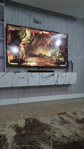 The video shows a TV screen displaying a Mortal Kombat game. The two characters are battling in a dark, jungle-like environment. The TV is mounted on a wall above a white cabinet with a marble pattern. The wall is decorated with vertical gray stripes. A rug with a brown and cream abstract pattern sits on the floor. A white PlayStation 5 console is visible next to the TV.