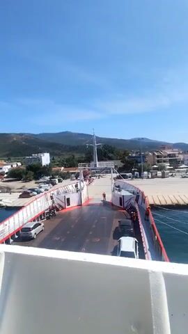 The video shows a view from the top deck of a ferry. The ferry is docked at a port. Cars are parked on the deck of the ferry. People are seen walking on the deck. The background shows a town with buildings and mountains. The sky is blue and clear. The video pans slightly to the right.