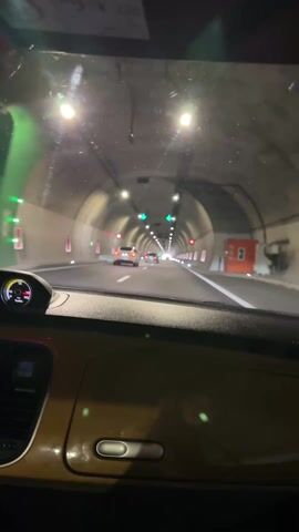 The video shows the view from inside a car driving through a well-lit tunnel. The car has a yellow dashboard.  Other cars are visible ahead in the tunnel. The tunnel has overhead lights, green arrows indicating directions, and red rectangular markers along the walls. The road has white lane markings.  The video is slightly shaky, suggesting it was filmed while driving.