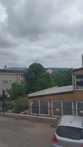 The video presents a slow, horizontal pan from right to left, showcasing a residential or mixed-use area under a cloudy sky in Meljine, Montenegro.

The scene opens with a view of several buildings, lush green trees, and distant hills. In the foreground on the right is a light yellow building with large, dark-framed windows, and beyond it, masts of boats or yachts are faintly visible, suggesting proximity to a marina.

As the camera pans left, a large, multi-story white building with red-tiled roofs, multiple balconies, and air conditioning units comes into full view. A satellite dish is mounted on its roof. In front of this building, a small grassy yard contains a car covered with a white tarp. A blue sign, partially obscured but clearly displaying "APARTMANI" in white text, is affixed to a fence near the building, likely advertising apartment rentals. A concrete path or square is visible in the immediate foreground.

Further to the left, a large, leafy green tree dominates the mid-ground, and bright pink flowering shrubs add a splash of color. Another building with a red roof is visible behind the foliage. The overall impression is one of a quiet, somewhat overgrown but developed area, typical of a coastal town. The overcast weather lends a muted tone to the landscape.