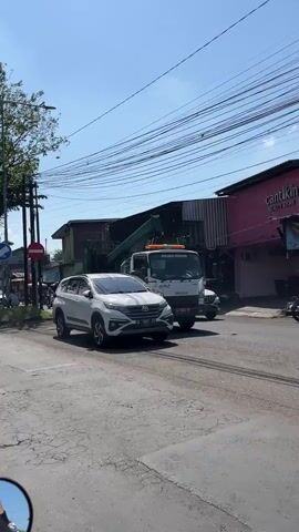 A white SUV is parked on a paved road. Behind the SUV, a green garbage truck is stopped. The truck has a large crane attached to its rear. A building with a pink facade is visible on the right side of the frame. Power lines crisscross the sky. The sky is clear and blue. The sun is shining. The scene appears to be a street in a town or city.