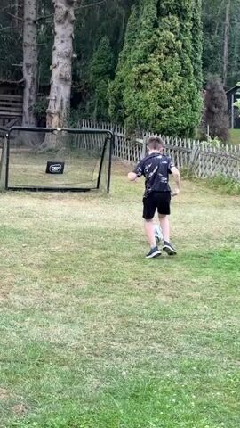 A young boy wearing a black t-shirt with a white pattern and black shorts is playing soccer in a grassy backyard. He is dribbling a white and light blue soccer ball and is facing a small black soccer net with a white logo on it. The backyard is surrounded by trees and a wooden fence. The boy takes a shot on goal and then retrieves the ball.