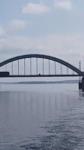 Here's a concise description of the video:

A gray arched bridge spans a wide, still river under a sky with light cloud cover. A train with multiple freight cars is moving across the bridge from left to right. The water reflects the sky and bridge. There's a transmission tower on one side of the bridge. Ripples in the water suggest the camera is on a moving boat.