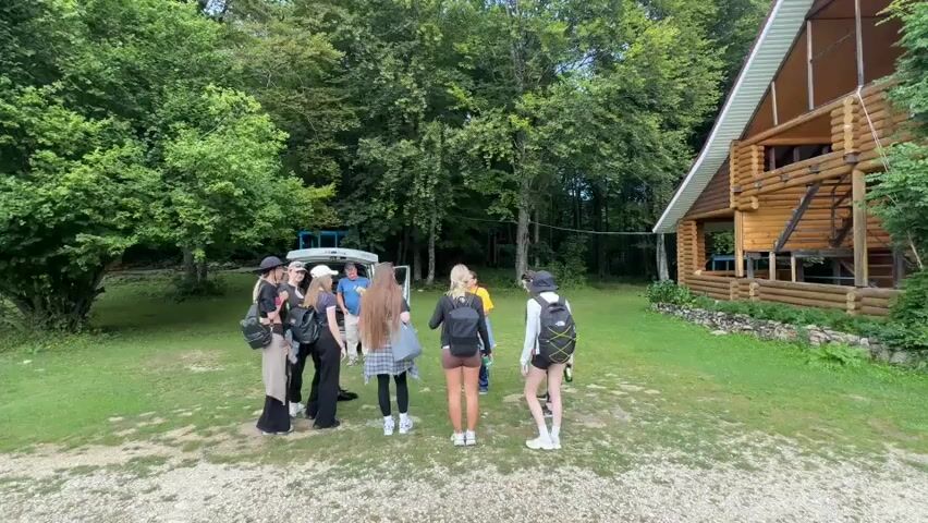 A group of nine young people, mostly female, stand in a clearing in front of a wooden lodge and a white van. The people are facing away from the camera, and are gathered around the open side door of the van. Some of the people are holding bags or backpacks. The clearing is surrounded by trees and grass. The weather appears to be overcast, but it is not raining.