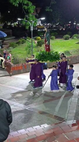 A group of people, mostly children and a few adults, are walking in procession across a brick-paved square at night. They are wearing matching purple robes and carrying wooden crosses and a wooden structure with green foliage on top. A statue of a woman stands in the background, illuminated by a streetlight. Other people are sitting or standing around the square, observing the procession. A sign in the background reads "YO ❤ TIUANA."