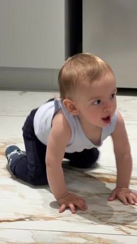 A blond baby wearing a white tank top and blue pants sits on a marbled tile floor in a room, possibly a kitchen, with white cabinets in the background. The baby is holding its hands in the air and looks to the right, seemingly reacting to music playing in the background.