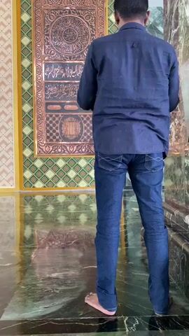 A person wearing a dark blue shirt and blue jeans is shown from behind performing Islamic prayer. They are in a room with a patterned wall and marble flooring. The person starts by standing, then bows and finally kneels with their forehead touching the floor. A decorative, ornate panel with Arabic calligraphy is visible on the wall behind them.