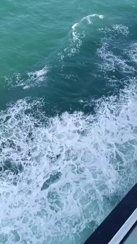 A high-angle shot shows the churning turquoise water of the ocean. White foam and waves rush towards the viewer on the right side of the frame, indicating movement. The water appears dark blue or green in the deeper sections. A dark, blurred railing is visible along the bottom right edge of the frame.