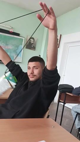 Here is a description of the video.

The video shows a young man sitting at a wooden school desk in a classroom. He is wearing a black hooded sweatshirt. With both arms raised, he dangles a juice or milk carton attached to a black string. The carton swings about. The classroom has light green walls and typical school desks. A map of Bulgaria hangs on the wall, along with a portrait of a man. Another student is sitting behind him. A black backpack is visible on a nearby desk.