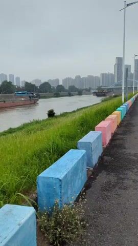 The camera pans along a paved riverside pathway on an overcast day. A row of vibrantly colored concrete blocks separates the path from the green grassy bank along the river. The blocks are painted in a rainbow of colors including blue, pink, yellow, and green. In the background, a wide river flows with cargo barges visible. A cityscape of tall buildings is seen in the distance. The air is still and the environment appears calm.