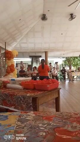 A person is standing with their back to the camera, facing an audience seated on chairs. The person is wearing an orange shirt and jeans. In front of the person, on a table covered with a colorful cloth, are two red plastic containers filled with red objects, possibly tomatoes. To the left of the table, there is a display of orange and yellow balloons. The environment appears to be an outdoor or semi-outdoor covered area with a ceiling and exposed beams. The audience consists of several adults sitting and listening to the speaker. There are also fans mounted on the ceiling.