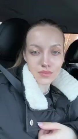 A woman in a black jacket with a white fur collar is sitting in a car. She has light skin, light eyes, and her hair is pulled back. She has light makeup on. She is wearing a seatbelt. She looks at the camera and then glances to the side.  She is wearing earrings and rings. The interior of the car is visible, including the headrest of the seat and a window. There are trees visible through the window.