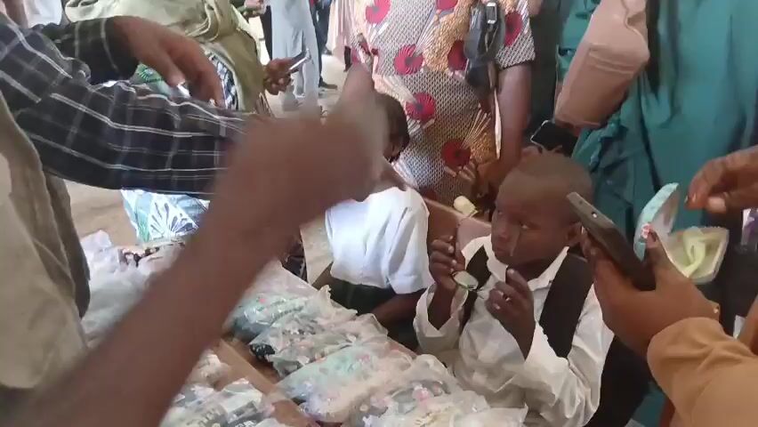 A group of people are gathered around a table with various items on it. In the foreground, two children, a girl with glasses and a boy, are seated at the table. The girl is wearing a white shirt and a green skirt, while the boy is in a white shirt and black shorts. They appear to be interacting with the items on the table, possibly making a purchase or receiving something. Several adults are standing around them, some holding cell phones. The environment appears to be indoors, possibly a marketplace or a school event. The items on the table are mostly small, packaged goods, some appearing to be snacks or candies. The lighting is bright, suggesting an open and well-lit space.
