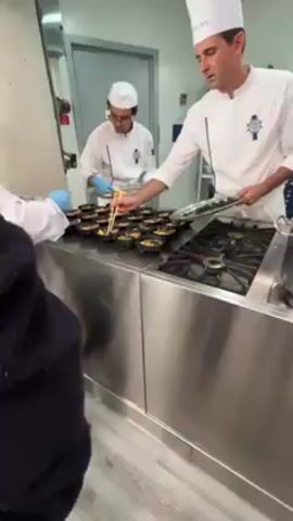 In a commercial kitchen, three chefs in white uniforms and blue gloves are preparing individual servings of a meal. The chefs are working on a stainless steel countertop next to a gas stove. Rows of small, black bowls are lined up on the countertop, each containing a similar arrangement of food: possibly salmon, spinach, and a half of a boiled egg. One chef is adding sliced hard-boiled eggs from a silver tray into each bowl. Another chef uses chopsticks to place green garnish into each bowl. The third chef pours a reddish sauce from a stainless steel bowl into the individual servings. The kitchen is brightly lit and appears clean and organized.