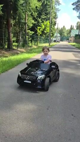 A young child, likely a toddler, is seated in a black toy Mercedes-Benz SUV and is in motion on a paved road or driveway. The child is wearing a light-colored t-shirt and appears to be steering the toy car. The background consists of a grassy area and trees on either side of the road, with a fence visible on the left. Houses and more trees are present in the distance. The lighting suggests it is daytime with sunlight.