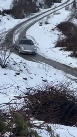 A silver Porsche sports car slowly drives up a snow-covered, narrow paved road. The sides of the road are covered in deep snow, with piles of brush and small trees lining the area. The road has tracks, showing other vehicles have already driven on it. The day appears overcast and the snow is lightly melting.