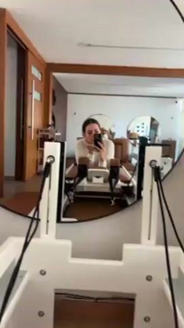A woman takes a photo of herself in a mirror while in a room with Pilates equipment. The mirror shows a view of the woman lying prone on a light brown Pilates reformer, wearing a white top and holding a black smartphone. Her hair is pulled back. The mirror's frame is dark brown. In the background, further into the room, are two more Pilates reformers and circular mirrors. The room has a light brown and white color scheme.  To the left is a partially visible wooden door. The camera view is of the lower part of another white Pilates reformer, showing ropes and hardware.