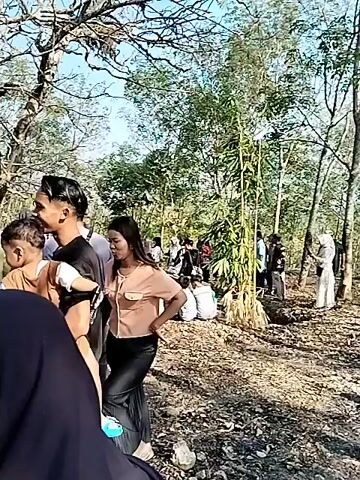 The video depicts a crowded outdoor scene with many people gathered. In the foreground, a man is holding a young child. To the right of them, a woman is partially visible. Behind them, a larger group of people are seen, some sitting and some standing, in what appears to be a gathering or event. The environment includes trees, some structures resembling stalls or small buildings, and a bright, possibly sunny, atmosphere. The overall impression is a busy social gathering in an open-air setting.