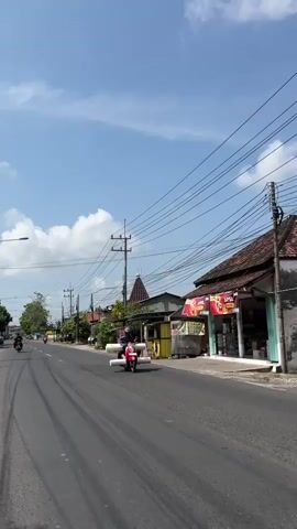 A person is riding a red scooter down a paved road. The scooter is carrying two large, white cylindrical objects strapped to the front. The person is wearing a dark shirt and a helmet. The road is lined with buildings and trees. There are power lines overhead. The sky is blue with some clouds. There are tire marks on the road.