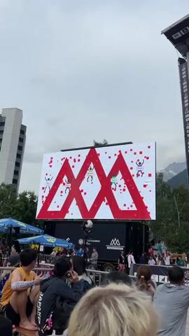 A large LED screen displays a climbing competition scoreboard. Three large red mountain peaks are outlined on the screen, with small red squares scattered around them. Silhouettes of climbers are positioned on or near the peaks, with their names above them. The names visible are Skip, Dani, Mathias, and Dani. The screen is mounted on the back of a black truck. In front of the screen, a crowd of people are watching. Some are standing, others are sitting. There are tents and umbrellas to the left of the screen. The environment appears to be outdoors with trees and mountains visible in the background. The sky is overcast.