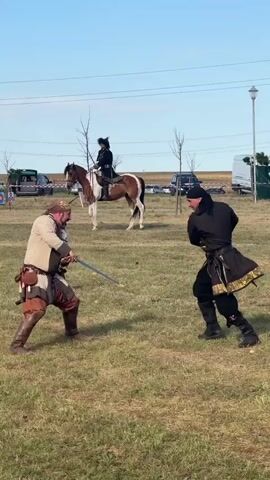 Two men are engaged in a staged sword fight in an outdoor grassy area. One man is wearing historical garb consisting of a light-colored tunic, brown breeches, and a head covering. He is holding a sword in his right hand and appears to be in a defensive stance. The other man is dressed in a dark, ornate tunic with gold trim and black pants, also holding a sword. He is in a more aggressive, forward-leaning stance, preparing to strike. In the background, a third person is riding a brown and white horse, wearing a dark hat. The environment is a open field with sparse trees and some vehicles and tents visible in the distance, under a clear blue sky. The overall impression is a historical reenactment or demonstration.