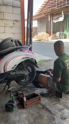 Here's a detailed description of the video:

A man is working on a motorcycle tire in what appears to be a repair shop or garage. He is squatting on a small block of wood next to a pink and white scooter with "Scoopy" written on it. The scooter is on its kickstand.

The man is wearing a green t-shirt with writing on the back and gray shorts, with green sandals. He is focused on handling a black tire. A wooden toolbox containing various tools is on the ground near him, along with other equipment like a green can.

Behind the scooter, there's a stack of tires leaning against a white brick wall. In the background, across the street, is a metal building, pallets stacked, and construction materials. A yellow truck passes by in the background. A second person also rides past on a motorcycle behind the truck. The scene is outdoors, and the lighting suggests it is daytime.