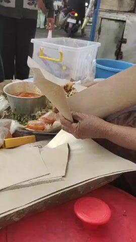 The video captures a close-up view of hands holding food wrapped in brown paper. One hand is shown holding a small plastic bag filled with orange sauce, seemingly drizzling it onto the food. Another hand is visible, also holding the brown paper. In the background, out of focus, there appears to be a market or street scene with people and stalls. A white plastic container and a red lid are also present in the foreground. The overall scene suggests a food vendor preparing or serving food.