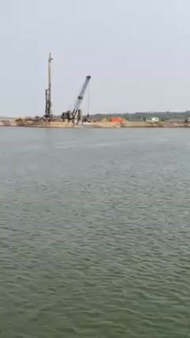 The video shows construction work on a body of water. The initial view shows construction equipment on a man-made island, including a crane and pile driver. Later shots reveal an excavator on the island, and a small boat carrying people is seen traveling in the water. The video then focuses on partially constructed bridge pillars rising from the water, with a construction crane and other equipment visible on the shore. Workers are seen on top of the pillars.