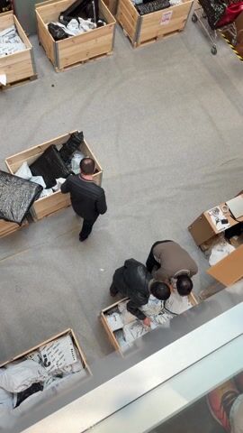 From a high angle, the video shows a section of a large, indoor commercial space, potentially a store or event hall. Several wooden crates are arranged on the floor, filled with items in plastic bags, some black and some white. Three men are actively rummaging through the crates. One man in a black jacket is walking between the crates. Two other men are bending over, looking at the items in one of the crates. Beyond the crates, there is a glimpse of a dining area with tables and chairs and a sign that says "MAISONS DU MONDE." A banner with the text "LOSTVS UNBOX THE FUN" is also visible. The floor is primarily a light gray color. The video is well-lit.