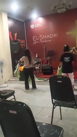 Two young women are dancing in front of a red wall with a gold cross and text that reads "EL-SHADAI Allah Maha Kuasa". The woman on the left is wearing a black t-shirt and light pink pants. The woman on the right is wearing a light-colored t-shirt and black shorts. Both women are holding long, green, leafy objects and waving them. There are musical instruments, including a keyboard and drums, on a stage behind them. Several black chairs are visible in the foreground. The overall environment appears to be a church or religious gathering space.