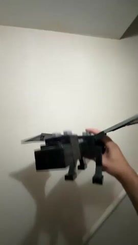A hand holds a black toy dragon with purple glowing eyes and a purple mouth. The dragon is made of blocks and has wings. The background is a plain wall. The dragon's head moves up and down as the hand moves it.