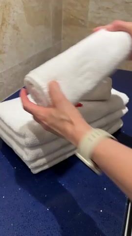 The video shows a person's hands arranging white towels. The towels are stacked on a dark blue surface in front of a tiled wall. The person places a rolled towel on top of a stack of folded towels. They then place two red rose petals on top of the rolled towel and another red rose petal on one of the folded towels. The person is wearing a watch on their left wrist.