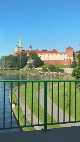 The video captures a scenic view of Krakow, Poland, with Wawel Castle prominently featured on a hill overlooking the Vistula River. The castle complex, with its red-tiled roofs and distinctive green-spired tower, is a central subject. The river flows in the foreground, reflecting the clear blue sky. A green railing runs across the bottom of the frame, suggesting the vantage point is from a bridge.

On the riverbank below the castle, there are several small boats and what appear to be outdoor seating areas with umbrellas, indicating a lively atmosphere, possibly a riverside cafe or market. Along the riverbank, a paved path winds through a grassy area, and a few people are visible enjoying the outdoors. One couple is seated on the grass near the path.

The overall scene is bathed in bright sunlight, suggesting it is daytime, likely mid-morning or afternoon, given the clear skies and shadows. The weather appears to be warm and pleasant. The presence of the historic castle, the river, and the recreational activities on the bank contribute to a feeling of peaceful exploration and enjoyment of a beautiful European city. No visible text is present in the video.