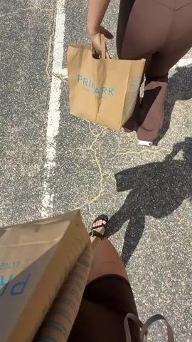 This is a first-person perspective video taken outdoors on a sunny day. The camera is pointed downwards, showing a person's legs and feet wearing black sandals and brown shorts. The person is carrying a brown paper bag with the Primark logo on it. The pavement has white painted lines and appears cracked. In the background, another person is walking away, also carrying a Primark bag. The scene then pans up, revealing a beach, the ocean, and a clear blue sky. The person holding the camera turns slightly to the right, showing their face and upper body wearing a brown crop top and holding another Primark bag. They are walking along a path next to the beach. In the further background, there are a few beach huts and a couple of people sitting on a bench.