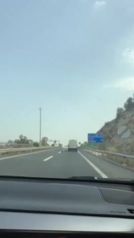This is a first-person perspective video taken from inside a car driving on a highway. The scene is set during the daytime under a hazy, bright sky, suggesting it could be late morning or afternoon with some atmospheric dust or pollution. The road is a multi-lane highway, with white lines marking the lanes. There is a concrete barrier on the right side of the road, and a grassy slope with some rocky outcroppings on the far right, indicating a hilly or mountainous terrain. Trees and vegetation are visible on the left side of the road.

Several vehicles are present on the highway. A white van is driving in the same direction as the camera car, ahead in the right lane. A silver car is visible in the left lane, passing the camera car. In the distance, there appear to be more vehicles and what might be overhead traffic signals or signs.

There are several road signs visible. On the right side of the highway, a blue sign with white text and symbols is partially visible, possibly indicating directions or services. Further down the road, a circular speed limit sign with "100" inside a red circle is visible on the right. Another similar speed limit sign, also showing "100," is visible on the left side of the road.

The overall mood is one of travel and transit. The weather appears clear but hazy, and the location suggests a highway drive in a region with some elevation, possibly in Southern Spain given the context of Lorca. No people are directly visible, only the implied presence of the driver and passengers within the camera car and occupants of other vehicles. The activity is simply driving on a highway.