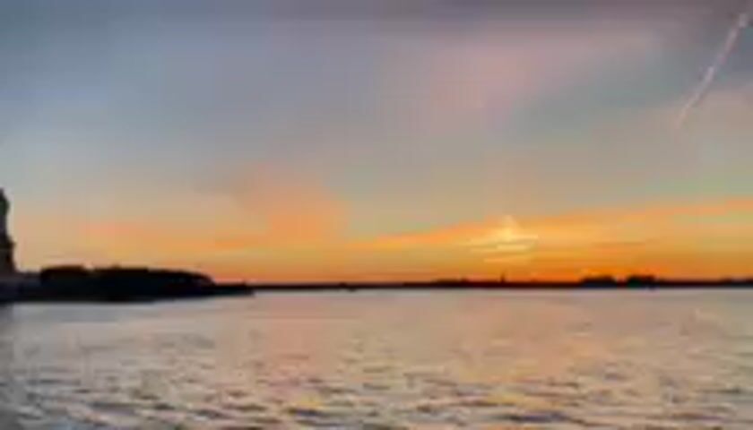 This media file shows a silhouetted view of the Statue of Liberty with a sunset in the background. The sky is painted with hues of orange, pink, and blue, with a contrail visible stretching across the upper right quadrant of the frame. The water in the foreground appears choppy, reflecting the colors of the sky. The overall scene suggests a serene and picturesque moment at dusk. No people are visible in the frame, and there is no visible text. The location is identified as Jersey City, United States.