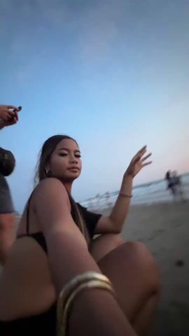 A young woman with her hair in a ponytail is sitting on a beach facing the ocean at sunset. She is wearing a black top with thin straps and has multiple gold bracelets on her arm. The camera is positioned close to her, focusing on her profile and arm. In the background, two people are standing near the water's edge. The sky is a mix of blue and orange hues from the setting sun.