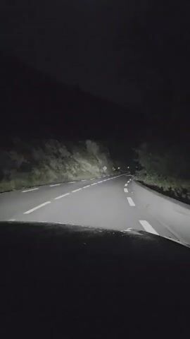 The video shows a view from a car driving at night on a winding road. The headlights illuminate the road ahead, revealing white lane markings and the dark slopes of a hillside on either side. There are a few streetlights visible in the distance along the road. The sky is dark and appears to be clear. The camera is positioned behind the dashboard, so the front of the car is visible in the foreground. The overall impression is of a peaceful night drive through a rural area.