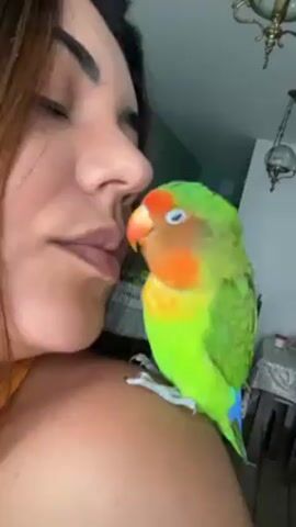 A woman with brown hair is holding a lovebird on her shoulder. She’s wearing an orange tank top. The lovebird is green with an orange face. The woman has her eyes closed. She gently touches the lovebird’s head with her finger. The lovebird’s beak touches her lips. A chandelier hangs in the background.