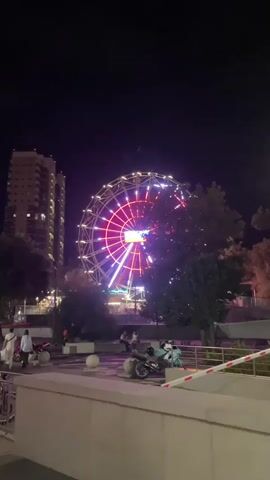 The video shows a Ferris wheel at night. The Ferris wheel is illuminated with red and blue lights and is rotating. In the foreground, there are several motorcycles parked. A few people are walking around the area. A tall building is visible in the background to the left of the Ferris wheel. The overall scene is an outdoor amusement area at night.
