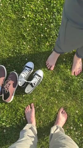 The video is an overhead shot of four pairs of feet on green grass. Two pairs of feet are bare, one pair is wearing brown sneakers, and another pair is wearing black Converse sneakers. The bare feet are positioned in the foreground and middle ground, with the shoes in the middle ground. The grass is short and there are small white flowers scattered throughout. The sun is shining, casting shadows on the grass. The scene appears to be outdoors on a sunny day.