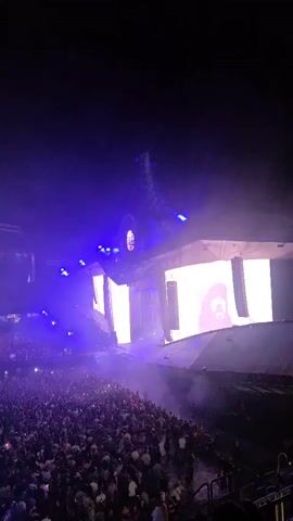 A wide shot captures a concert crowd in a stadium. The audience is packed together in the foreground, facing a large stage. The stage is illuminated with bright white lights and filled with fog, creating a dramatic atmosphere. A large screen on the stage displays a colorful, abstract image. In the background, more stadium seating is visible, also filled with people. The overall scene suggests a high-energy musical performance.