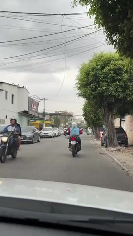 The video shows a street scene in what appears to be a city. There are several vehicles on the road, including motorcycles and cars. The motorcycles are traveling in the same direction as the camera, and there are also cars in front of and beside the motorcycles. The road is paved and there are buildings and trees on either side of the street. The sky is cloudy. The video appears to be shot from inside a car, looking out through the windshield. The timestamp on the video indicates it was recorded in 2023.